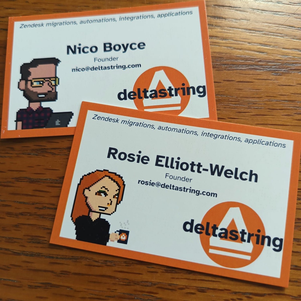 Two Deltastring business cards on a wooden surface, each with a thick solid orange border, white centre, the Deltastring logo, a pixel-art avatar, and the founder's name and email: Nico Boyce and Rosie Elliott-Welch.