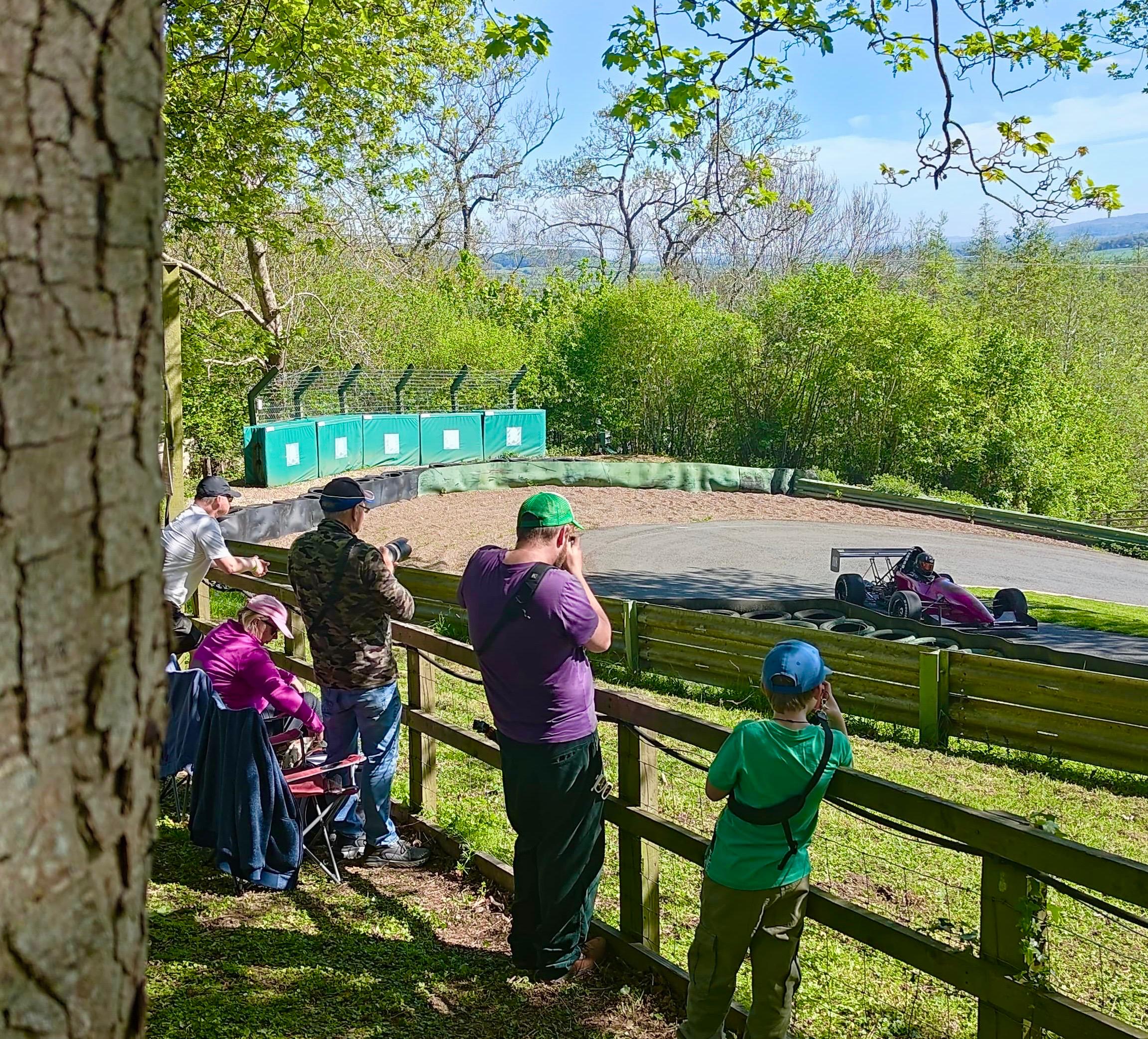 Photographers gathered at the fence of a motorsport circuit, karts visible on the track beyond