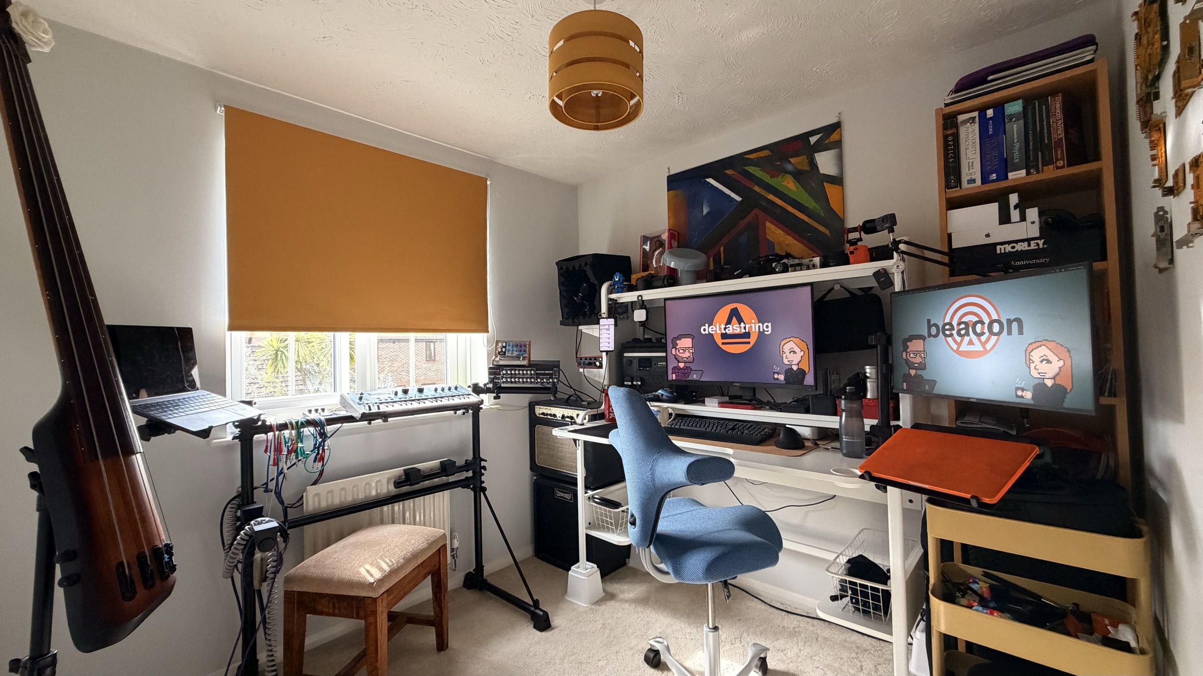 Wide-angle photo of Nico's home office. A double bass leans against the left wall next to a window with an orange roller blind. A keyboard stand holds a Novation synth. The centre desk has two monitors displaying the Deltastring and Beacon brands, a microphone on a boom arm, and a blue HÅG Capisco chair. A bookshelf on the right holds cameras, books, and trinkets. A yellow storage trolley sits in the bottom right corner.