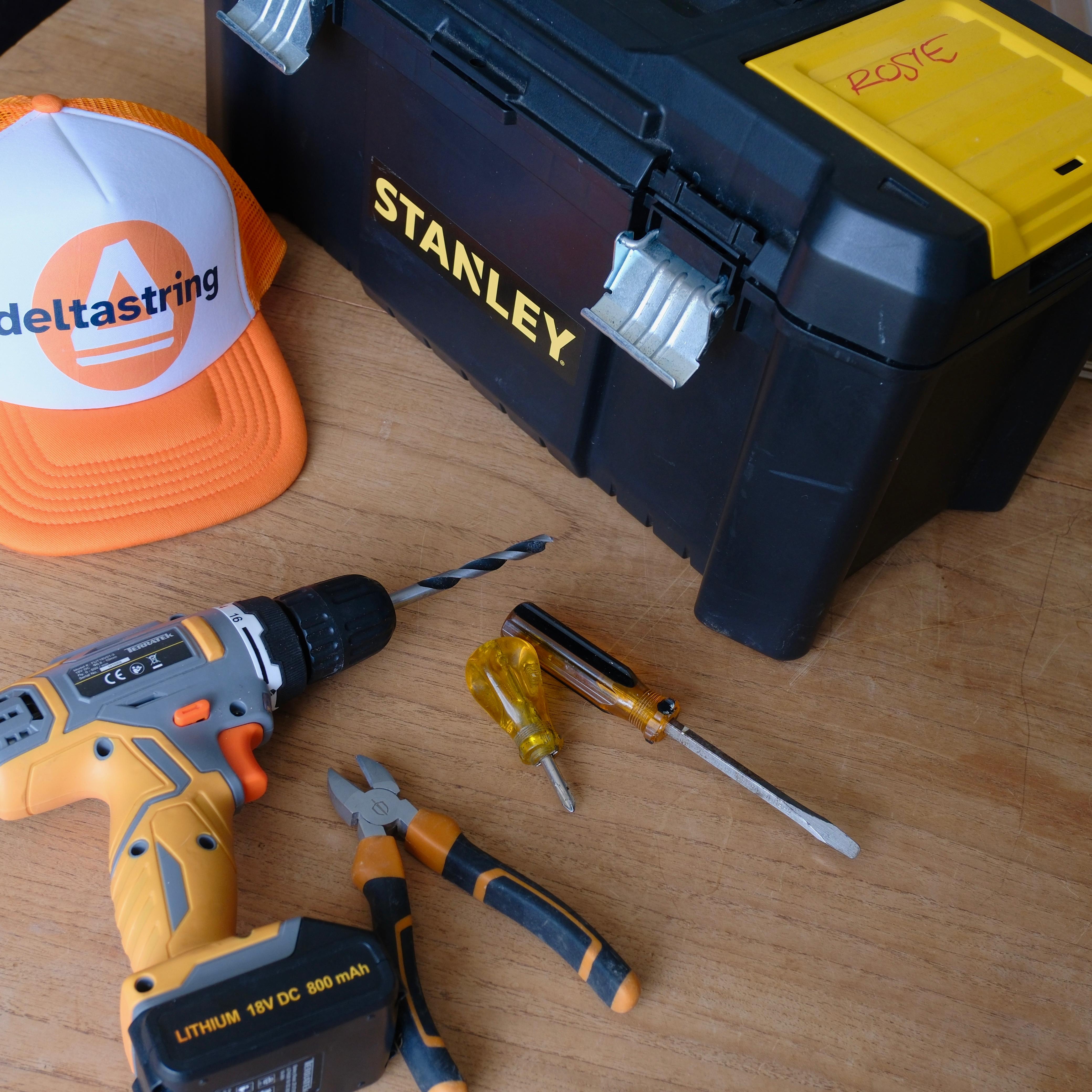 Rosie's Stanley toolbox marked ROSIE, a Deltastring cap, a cordless drill, pliers and a screwdriver laid out on a workbench